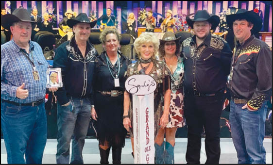 A group of seven adults wearing cowboy hats and western-style clothing stand together indoors around a tall sign that reads “Grand Ole Opry." Description generated by AI