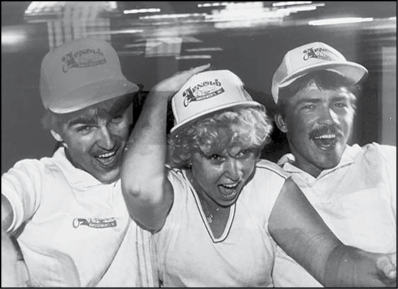 Three adults wearing matching hats and shirts are smiling and posing closely together in a lively setting. Description generated by AI