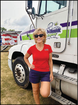 A person wearing sunglasses, a red shirt, and dark shorts is standing with one leg raised and leaning against a large white truck with green and purple stripes and the word “Schmidt" on the door. Description generated by AI