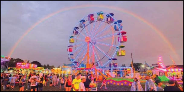 A brightly lit Ferris wheel stands in the center of a lively fairground filled with people under a sky displaying a vibrant rainbow. Description generated by AI