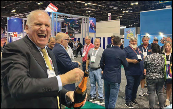 A person in a suit is smiling and opening a bottle of champagne while a group of people stand and talk in a convention or event setting. Description generated by AI